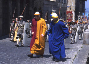 Men in medieval dress, shield and spear and long, blue, and red and orange tunics parade through an Arezzo street as part of the jousting festival.