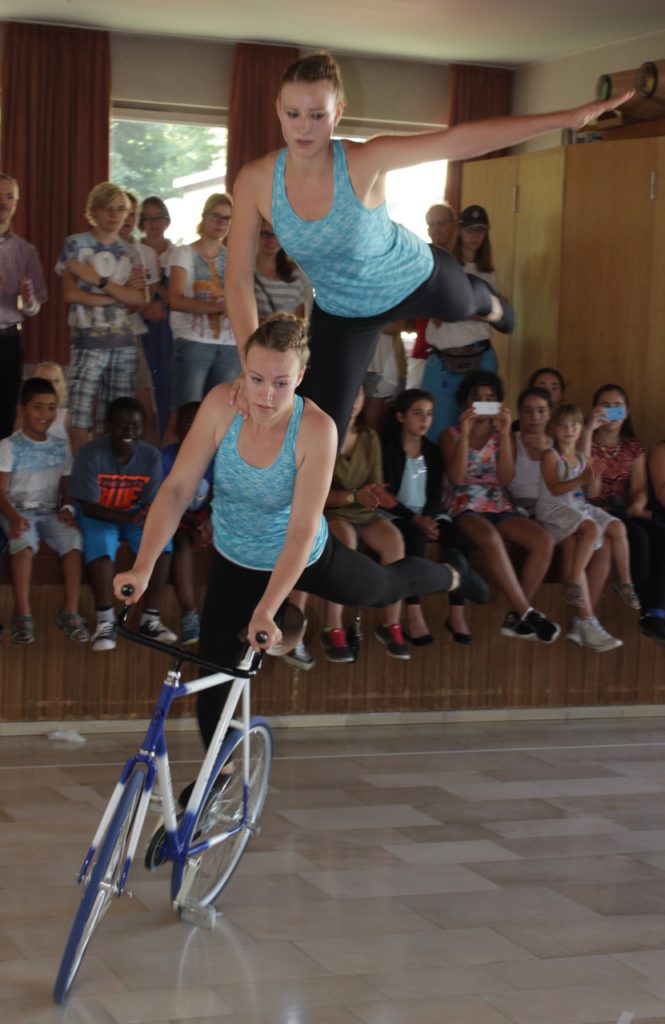 Two gymnasts riding the same bike. One standing on 1 leg with 1 arm in the air.