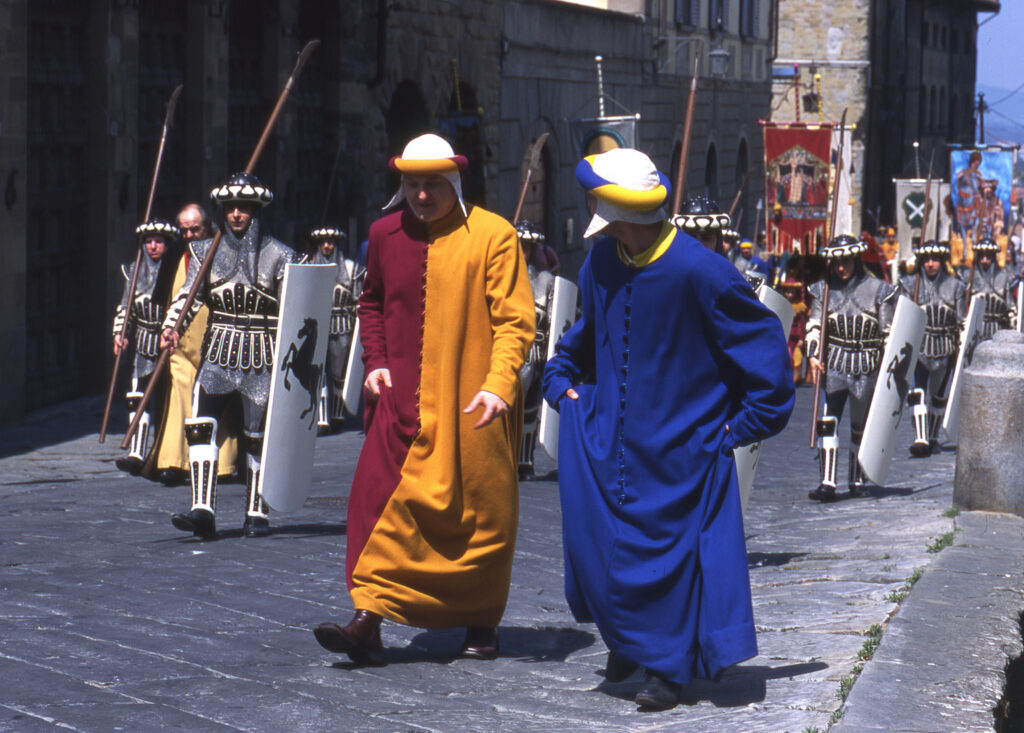 Men in red and gold and blue medieval woolen tunics accompanied by shield and spear bearing knights