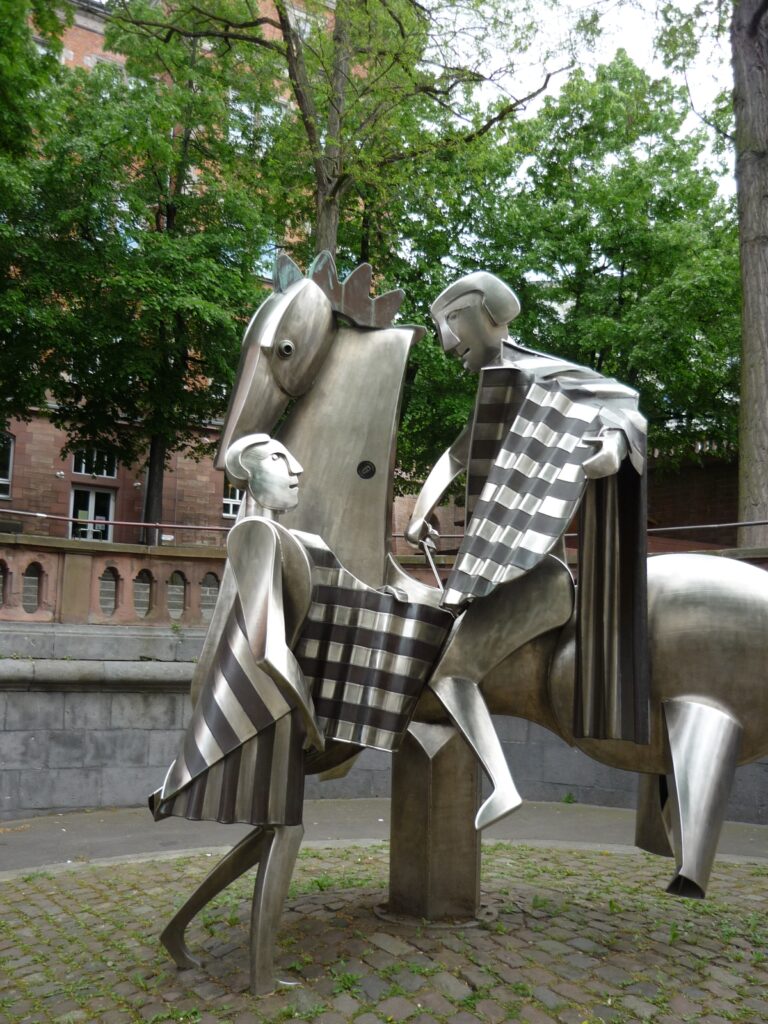 Metal sculpture of modernistic man on horseback dividing his patterned metal cloak with man on the ground.