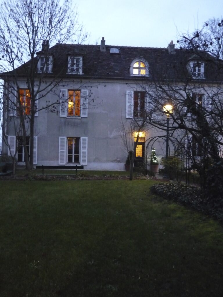 House with lit up windows as night falls in winter. Bare trees, green grass, pale blue sky.