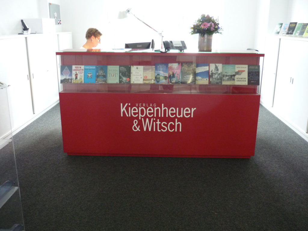 bright red front desk with name of publisher in white and a row of books in a built in shelf