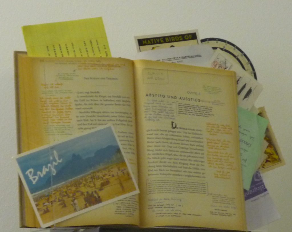 Printed book with marginal notes in two ink colors and formatted handwritten lists, postcards and other papers tucked in strategically.