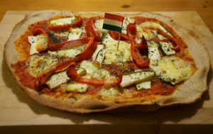 pizza baked on a stone with pepper and zucchini length-wise slices, mozzarella cheese, tomato sauce and an Italian flag toothpick in the center.