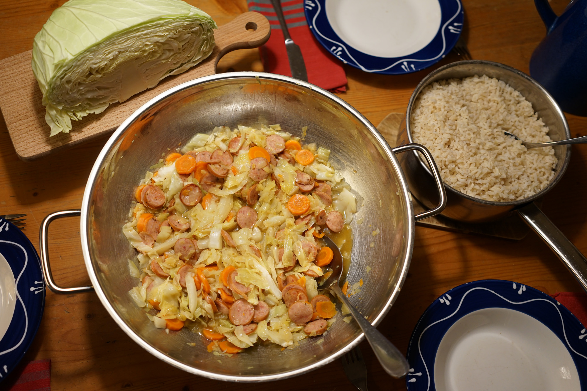 wok with sliced cabbage, carrots and kielbasa sausage next to a pot of brown rice and a flat variety cabbage cut in half