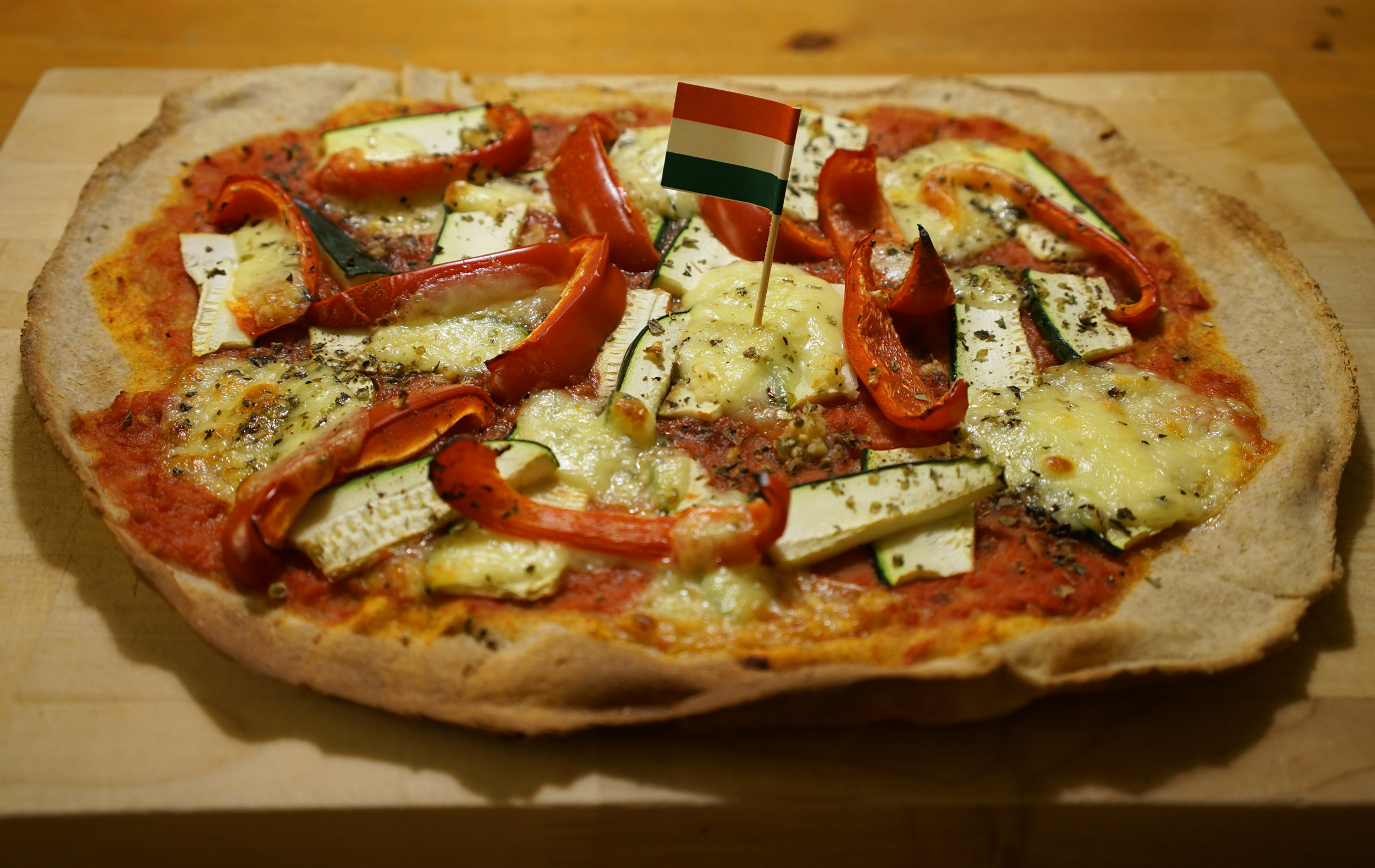 pizza baked on a stone with pepper and zucchini length-wise slices, mozzarella cheese, tomato sauce and an Italian flag toothpick in the center.